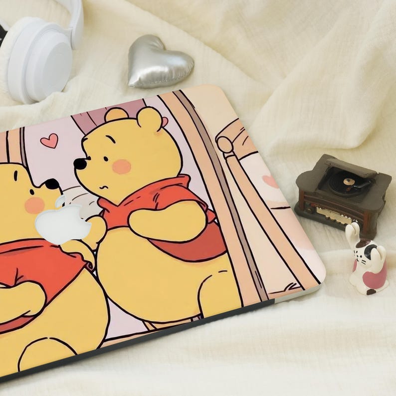May include: A laptop skin featuring a cartoon illustration of Winnie the Pooh and a friend, both in red tops. The skin has a light pink background with a heart and a window design. A small record player and a rabbit figurine are also visible.