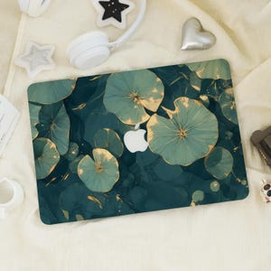 May include: A laptop skin featuring a teal and gold floral design. The design includes large, stylized lily pads with gold accents against a dark teal background. The Apple logo is visible. The skin is rectangular and designed to fit a laptop.