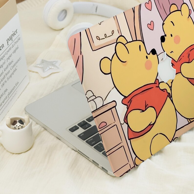 May include: A laptop featuring a Winnie the Pooh design on its cover. The cartoon shows two yellow bears in red shirts. The laptop sits on a white surface, alongside white headphones and a small ceramic cup.