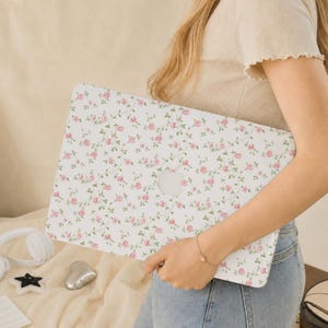 Puede incluir: Una funda rígida blanca para portátil con un patrón repetitivo de pequeñas rosas rosas con tallos y hojas verdes. La funda tiene un recorte en el centro para dejar ver el logotipo de Apple plateado en la tapa del portátil bajo la cubierta.