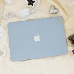 May include: A light blue laptop with a silver Apple logo on the lid. The device is surrounded by white headphones, a silver heart-shaped object, a white star-shaped ornament, a black star-shaped ornament, and a small white ceramic cup.