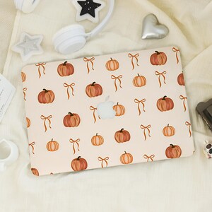 May include: A laptop cover featuring a repeating pattern of orange pumpkins and orange ribbons on a light cream background. The design includes a central cutout for the Apple logo, showcasing a clean, organized arrangement of seasonal motifs.
