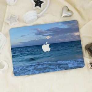 Puede incluir: Una funda para portátil con un diseño de paisaje marino, que muestra agua azul del océano, olas blancas y un cielo nublado. El logotipo de Apple es visible. La funda está sobre una superficie blanca con otros accesorios.