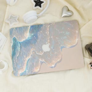 Può includere: Un laptop con una custodia perlata con un design astratto e fluido nei toni del blu, bianco e oro. Il logo Apple è visibile sulla custodia. Il laptop è circondato da accessori bianchi.