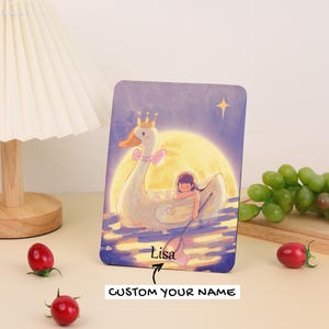 May include: A rectangular card featuring a whimsical illustration of a swan with a crown and a girl in a boat. The background includes a full moon and a star. The text "Lisa" and "CUSTOM YOUR NAME" are also visible.
