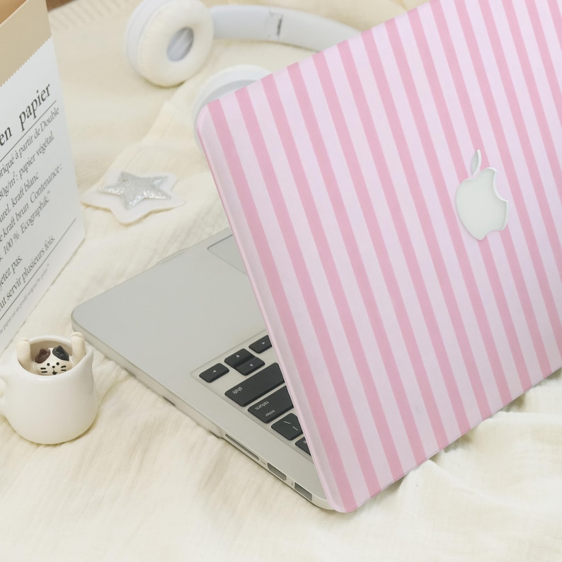K&ouml;nnte beinhalten: Ein silberner Laptop mit einer rosa-wei&szlig; gestreiften H&uuml;lle. Die H&uuml;lle hat ein wei&szlig;es Apple-Logo. Der Laptop ist ge&ouml;ffnet und zeigt die Tastatur. Eine wei&szlig;e sternf&ouml;rmige Dekoration und eine kleine Keramik-Katzen-Tasse sind ebenfalls zu sehen.