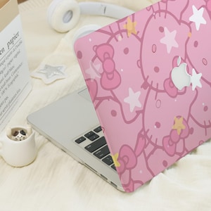May include: Pink laptop case featuring a repeating pattern of cartoon characters, bows, and stars. The case is on a silver laptop, with a white Apple logo visible. A white paper bag with text and a small ceramic cup are in the background.