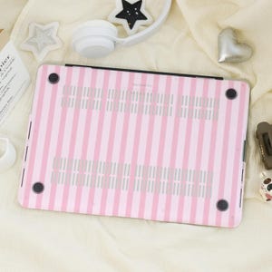 K&ouml;nnte beinhalten: Eine rosa-wei&szlig; gestreifte Laptop-H&uuml;lle. Die H&uuml;lle hat ein vertikales Streifenmuster und ist f&uuml;r einen Laptop konzipiert. Sie hat vier schwarze Gummif&uuml;&szlig;e und einen schwarzen Rand.