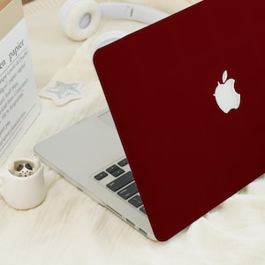 May include: A burgundy laptop with a white Apple logo is partially open, revealing the keyboard. The laptop is on a white surface with a paper bag, headphones, and a small ceramic cup with a cat design.