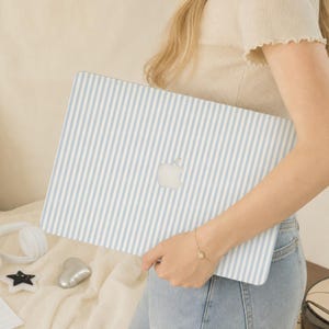 May include: A laptop skin featuring a pattern of thin, vertical light blue and white stripes. The skin is applied to the exterior of a laptop, with the central Apple logo cutout clearly visible against the striped design on the device cover.