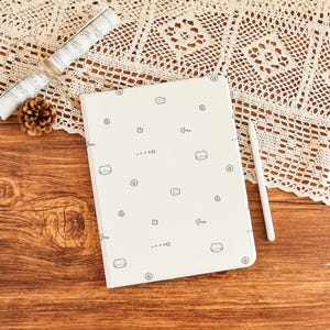 Puede incluir: Un cuaderno color crema con un patrón de caras de gato, espinas de pescado y espirales. Un lápiz óptico blanco descansa junto al cuaderno sobre una superficie de madera. También hay notas musicales decorativas y una piña.