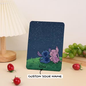 Peut inclure: Un objet décoratif rectangulaire bleu foncé représentant un ciel étoilé et les personnages de dessins animés Stitch et Angel. Les personnages sont assis sur de l'herbe verte. Le texte "Lisa" et "CUSTOM YOUR NAME" sont également visibles.