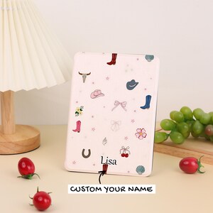 May include: A light pink e-reader case featuring a pattern of small illustrations including cowboy boots, hats, bows, flowers, cherries, a horseshoe, and a bull skull. The name Lisa is printed in black text above a small arrow pointing to the bottom.