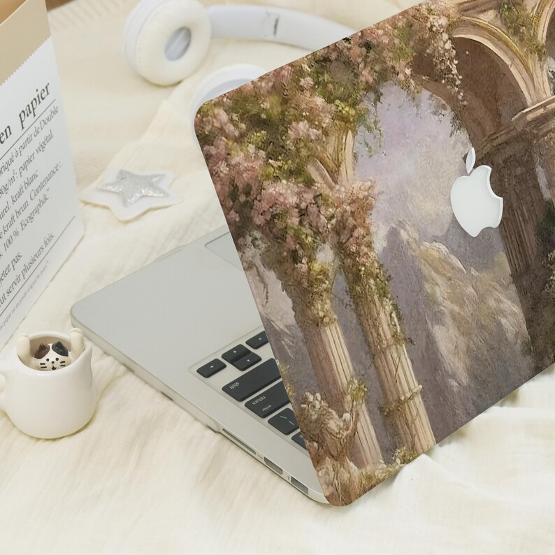 May include: A laptop with a decorative case featuring a classical architectural design with columns, arches, and floral accents. The case has a white Apple logo cutout. Other items include white headphones, a paper bag, and a small ceramic cup.