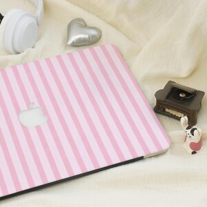 K&ouml;nnte beinhalten: Ein Laptop mit rosa und wei&szlig;en Streifen und dem Apple-Logo. Der Laptop liegt auf einer wei&szlig;en Oberfl&auml;che mit einem silbernen Herz, einem kleinen dekorativen Plattenspieler und einer Hasenfigur.