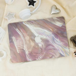 Pode incluir: Um laptop com uma capa perolada e iridescente com um design abstrato e fluido em tons de rosa, roxo e branco. O logotipo da Apple é visível. O laptop é cercado por acessórios brancos.