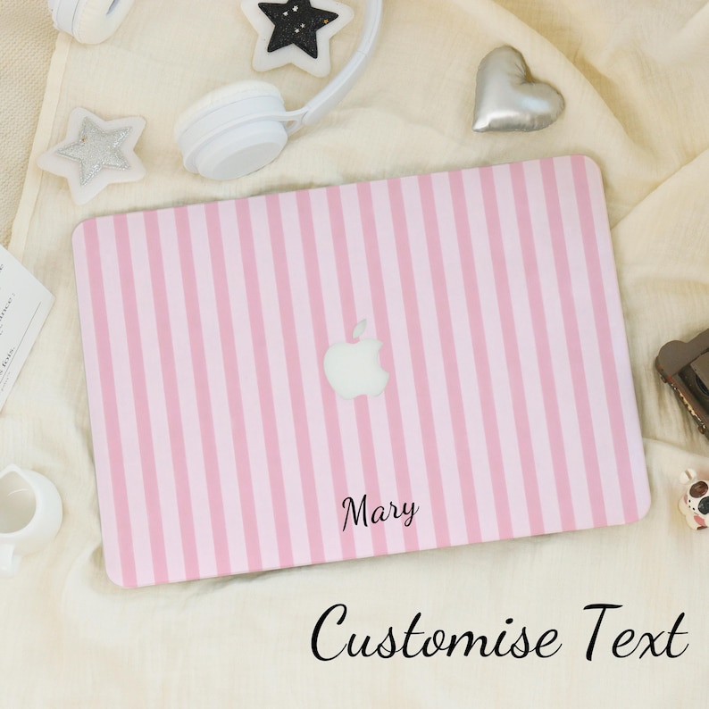K&ouml;nnte beinhalten: Eine rosa-wei&szlig; gestreifte Laptop-H&uuml;lle mit dem Apple-Logo. Der Name "Mary" ist in schwarzer Schrift gedruckt. Die Worte "Customise Text" sind darunter in Kursivschrift geschrieben. Wei&szlig;e Kopfh&ouml;rer und dekorative Sterne umgeben die H&uuml;lle.