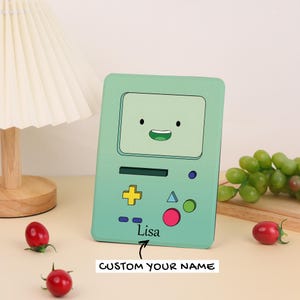 May include: A light green rectangular case with a cartoon face design. The case has a yellow cross button, a pink circle, and the name "Lisa" printed on it. The text "CUSTOM YOUR NAME" is also visible.