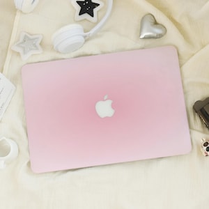 Puede incluir: Un portátil rosa claro con el logotipo de Apple en el centro. El portátil está rodeado de auriculares blancos, decoraciones en forma de estrella y un corazón plateado sobre una superficie crema.