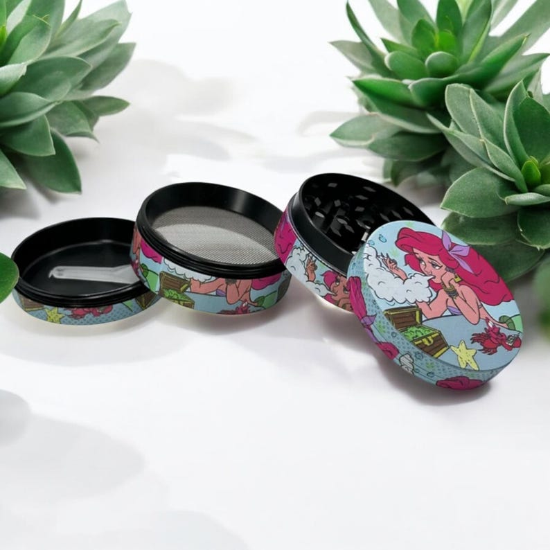 May include: An open, disassembled grinder with a colorful design featuring a cartoon mermaid. The grinder is black and has a mesh screen. The design includes a mermaid, treasure chest, and other aquatic elements.