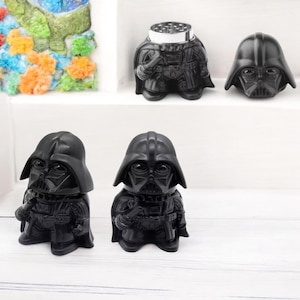 May include: Black Darth Vader grinder with a silver top. The grinder is shaped like Darth Vader's head and body. There are three grinders in the image.