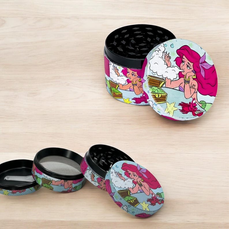 May include: A set of three-piece herb grinders with a black interior and a colorful exterior featuring a cartoon mermaid with pink hair. The grinder has a lid, a grinding chamber, and a bottom chamber.