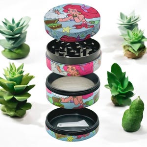 May include: A four-piece herb grinder with a colorful design featuring a mermaid with red hair. The grinder is disassembled, showing the different components, including a mesh screen and a small scoop. The background includes several small succulent plants.
