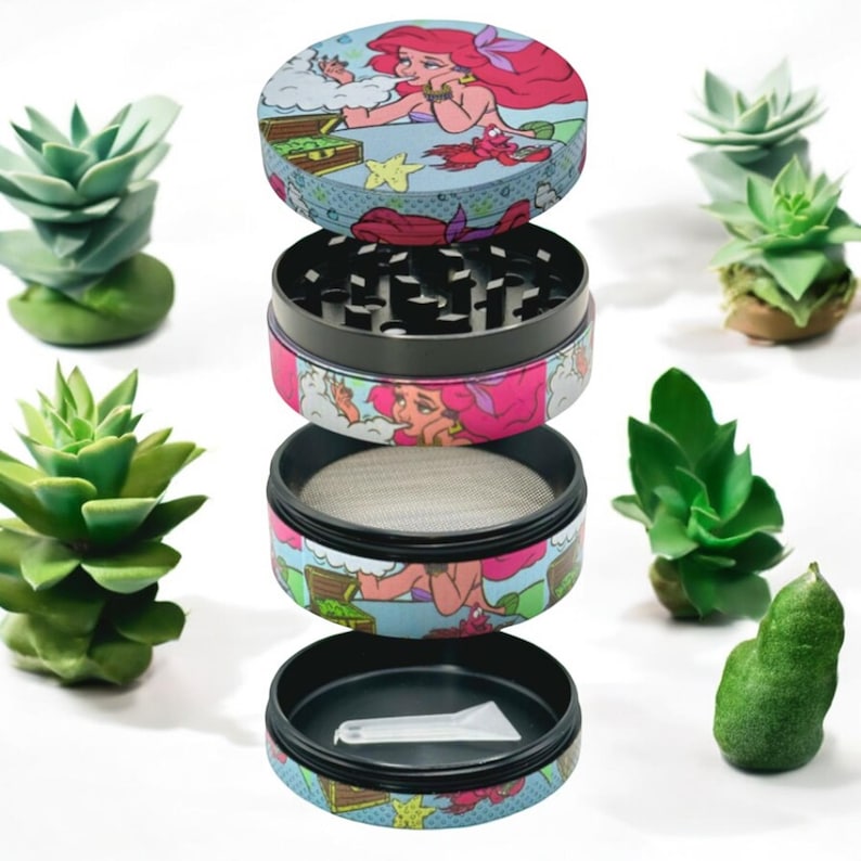 May include: A four-piece herb grinder with a colorful design featuring a mermaid with red hair. The grinder is disassembled, showing the different components, including a mesh screen and a small scoop. The background includes several small succulent plants.