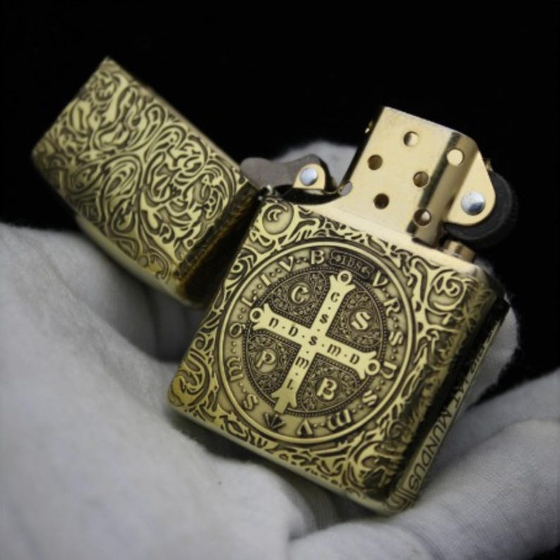 May include: A gold-toned lighter with an intricate, engraved design. The lighter features a cross with the inscription "G.S.S. D-DS-M-D. P.B.L.S.M.D.U.S.MUNDU".