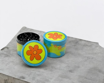 May include: Two blue and green metal grinders with a flower design. The top of the grinder has a large orange flower with a yellow center. The bottom of the grinder has a smaller orange flower with a yellow center.