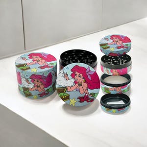 May include: A multi-piece herb grinder with a cartoon illustration of a mermaid with pink hair. The grinder is cylindrical with a black grinding surface and a light blue background with a treasure chest and starfish.