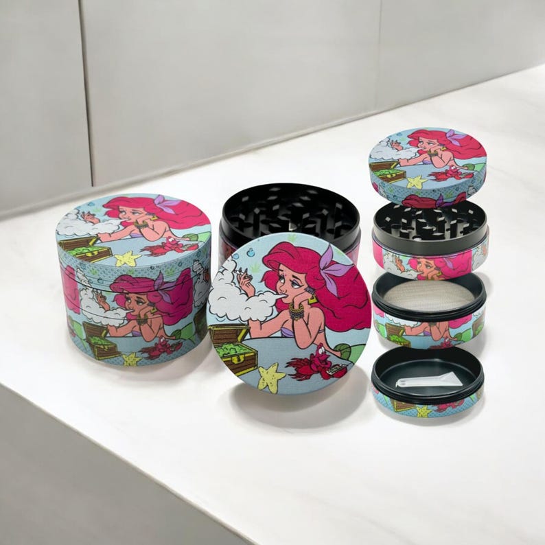 May include: A multi-piece herb grinder with a cartoon illustration of a mermaid with pink hair. The grinder is cylindrical with a black grinding surface and a light blue background with a treasure chest and starfish.