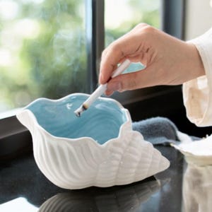 May include: White ceramic seashell ashtray with a light blue interior. A cigarette is being extinguished in the ashtray.