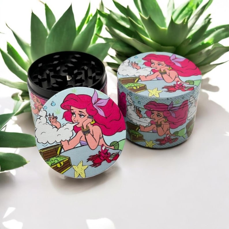 May include: A black and silver herb grinder with a lid featuring a cartoon mermaid with pink hair, a treasure chest, and a red crab. The grinder is cylindrical and has a textured top.