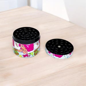 May include: A two-piece grinder with a black top and a colorful wrap featuring a cartoon mermaid. The grinder is open, showing the grinding teeth. The wrap has pink, green, and white accents.