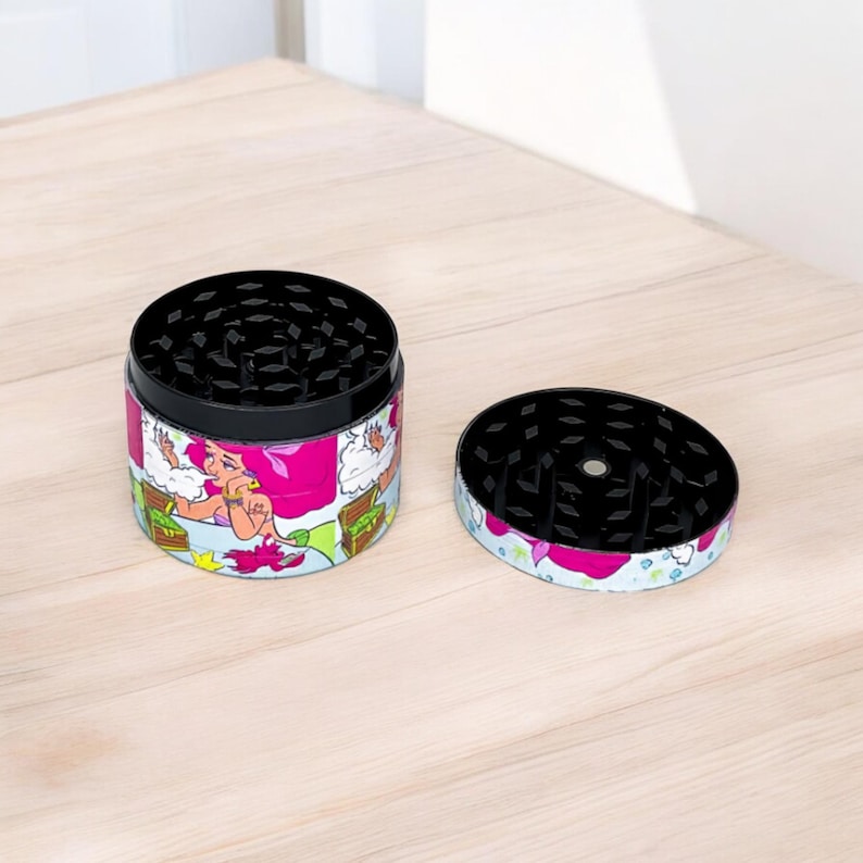 May include: A two-piece grinder with a black top and a colorful wrap featuring a cartoon mermaid. The grinder is open, showing the grinding teeth. The wrap has pink, green, and white accents.