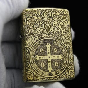 May include: A gold-toned lighter with an intricate, engraved design. The design features a cross with the words "S.D.S.M.D" and "P.M.B." around it. The lighter is surrounded by a floral pattern.