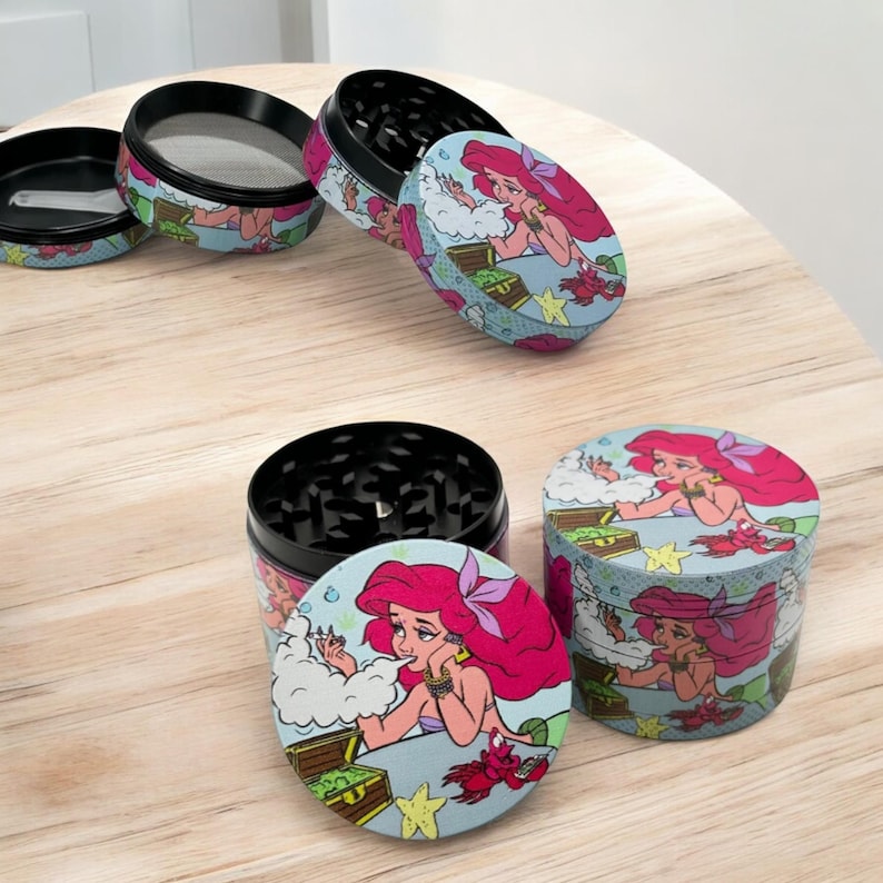 May include: A set of four-piece herb grinders with a colorful cartoon design. The grinders feature a pink-haired mermaid illustration on a light blue background with a treasure chest and crab. The grinders are black and silver.