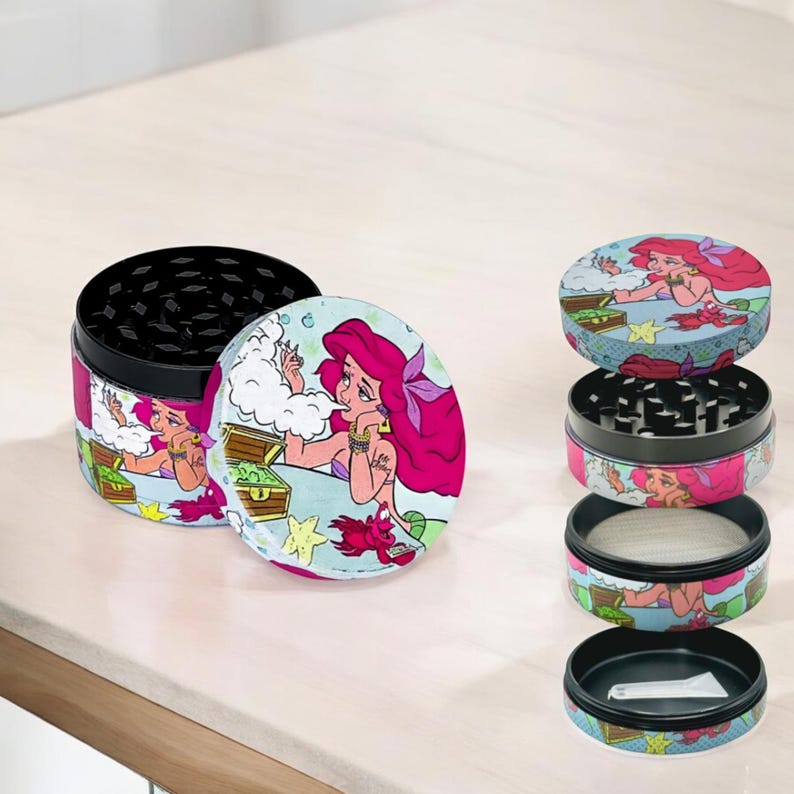 May include: A multi-piece grinder with a cartoon mermaid design. The grinder features a black interior with a mesh screen and a lid with a colorful illustration of a mermaid with pink hair. The grinder is designed for herbs and spices.