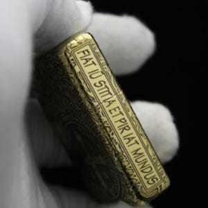 May include: A gold-colored lighter with an intricate engraved design. The lighter has a rectangular shape and features the Latin phrase "Fiat lux sitta et pir iat mundus" engraved on the side.