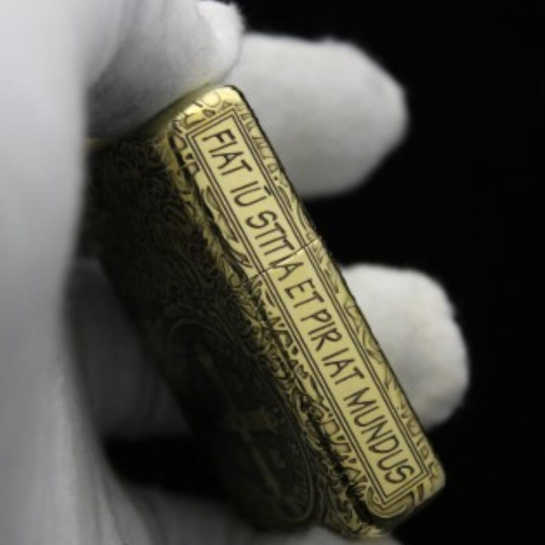 May include: A gold-colored lighter with an intricate engraved design. The lighter has a rectangular shape and features the Latin phrase "Fiat lux sitta et pir iat mundus" engraved on the side.