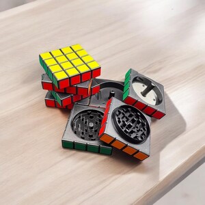 Puede incluir: Un molinillo negro y plateado con forma de cubo de Rubik. El molinillo tiene un diseño rojo, amarillo y verde. El molinillo está hecho de metal y tiene una tapa extraíble.