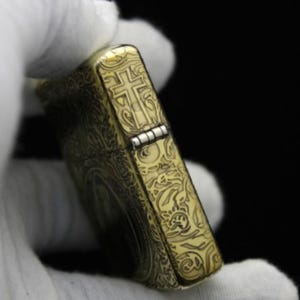 May include: A gold-toned lighter with an intricate engraved design featuring a cross and floral patterns. The lighter has a silver hinge.