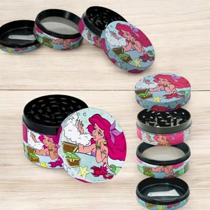 May include: Several multi-piece herb grinders with a cartoon illustration of a mermaid with pink hair. The grinders have a black top and bottom, with a colorful design on the sides and lids. The design includes a treasure chest and sea creatures.