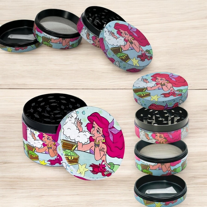 May include: Several multi-piece herb grinders with a cartoon illustration of a mermaid with pink hair. The grinders have a black top and bottom, with a colorful design on the sides and lids. The design includes a treasure chest and sea creatures.