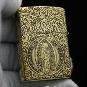 May include: A gold-colored lighter with an intricate engraved design featuring a religious figure and Latin text. The lighter has a detailed floral pattern around the edges.