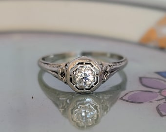 May include: A silver-toned metal ring featuring a single round brilliant-cut clear stone set within an octagonal frame. The band displays intricate engraved filigree detailing along the shoulders, leading to a polished, slender shank.
