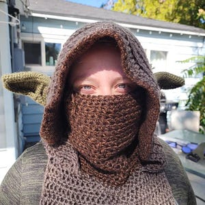May include: A brown and green crocheted hooded scarf with ear flaps. The scarf is worn by a person, but the person's face is covered by the scarf.