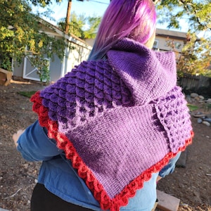 Puede incluir: Un chal de ganchillo morado con capucha que presenta un patrón de escamas texturizado en la sección superior y un diseño de punto sólido en la parte inferior, acabado con un borde festoneado rojo a lo largo de todo el contorno.