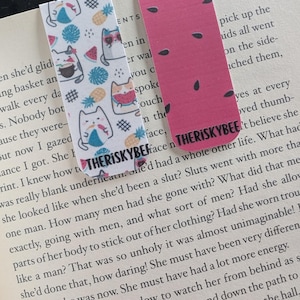 Könnte beinhalten: Zwei rechteckige Lesezeichen. Eines hat einen weißen Hintergrund mit Katzenillustrationen, Ananas und Wassermelonenstücken. Das andere ist rosa mit schwarzen Wassermelonenkernen. Beide haben den Text "THERISKYBEE".